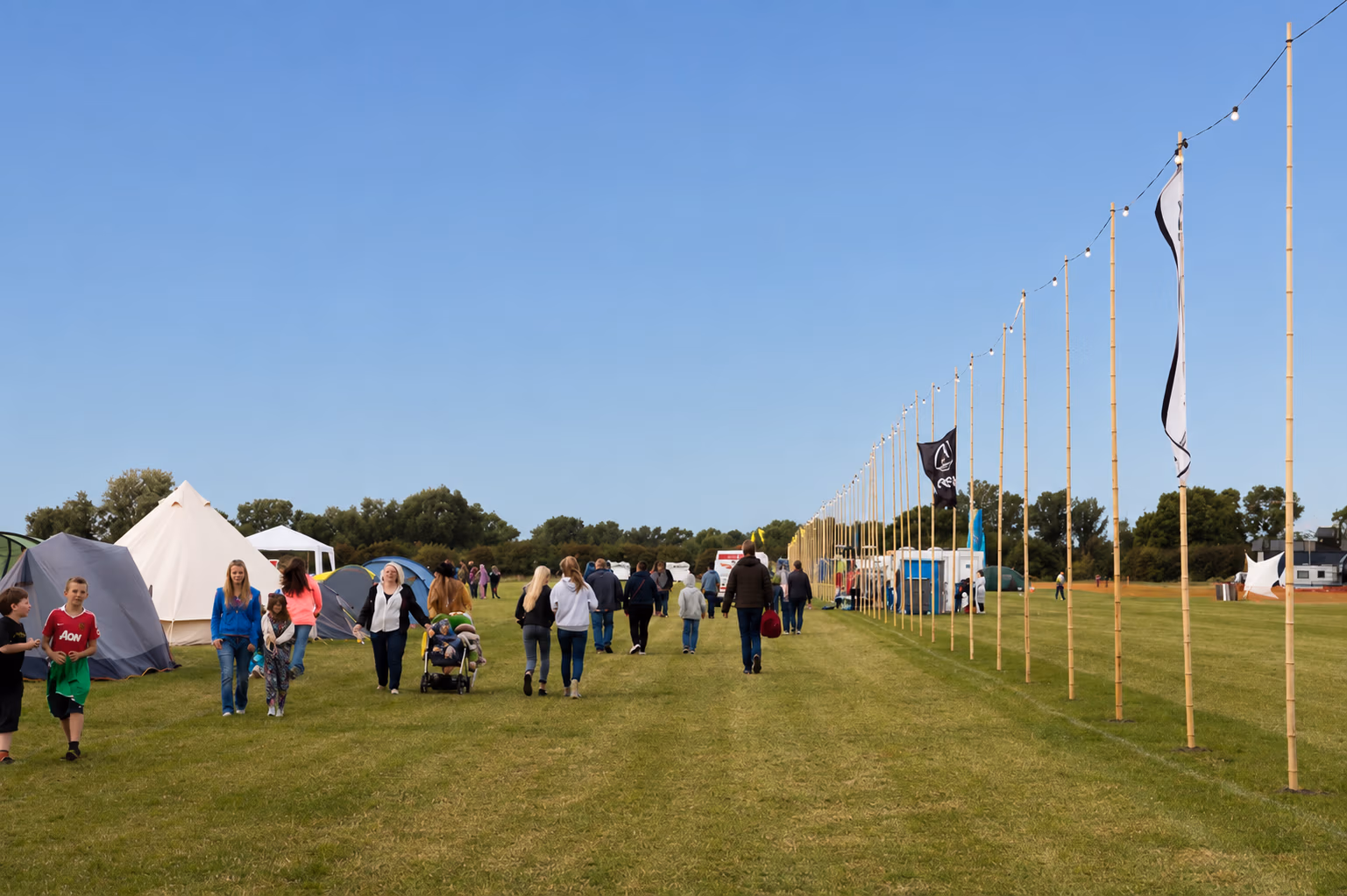 Campsite walkway with LodeStar Festival-goers arriving and settling in the festival near Cambridge. Rows of bamboos carry the festoon lights.